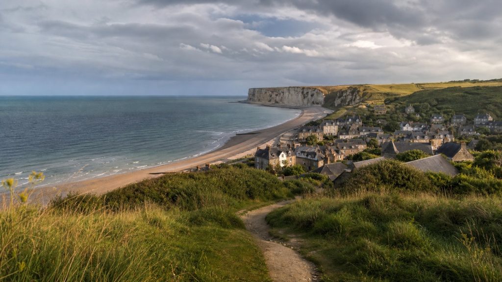 Vue panoramique de la côte de Basse-Normandie avec plage, falaises blanches, village aux toits d’ardoise et sentier côtier sous un ciel nuageux.