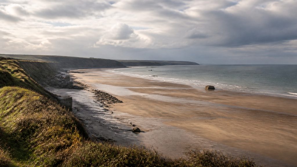 Vue panoramique d’une plage du Débarquement en Normandie à marée basse, bordée de falaises verdoyantes sous un ciel nuageux.
